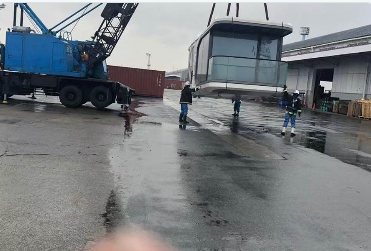 Crane lifting a capsule cabin at a wet industrial yard