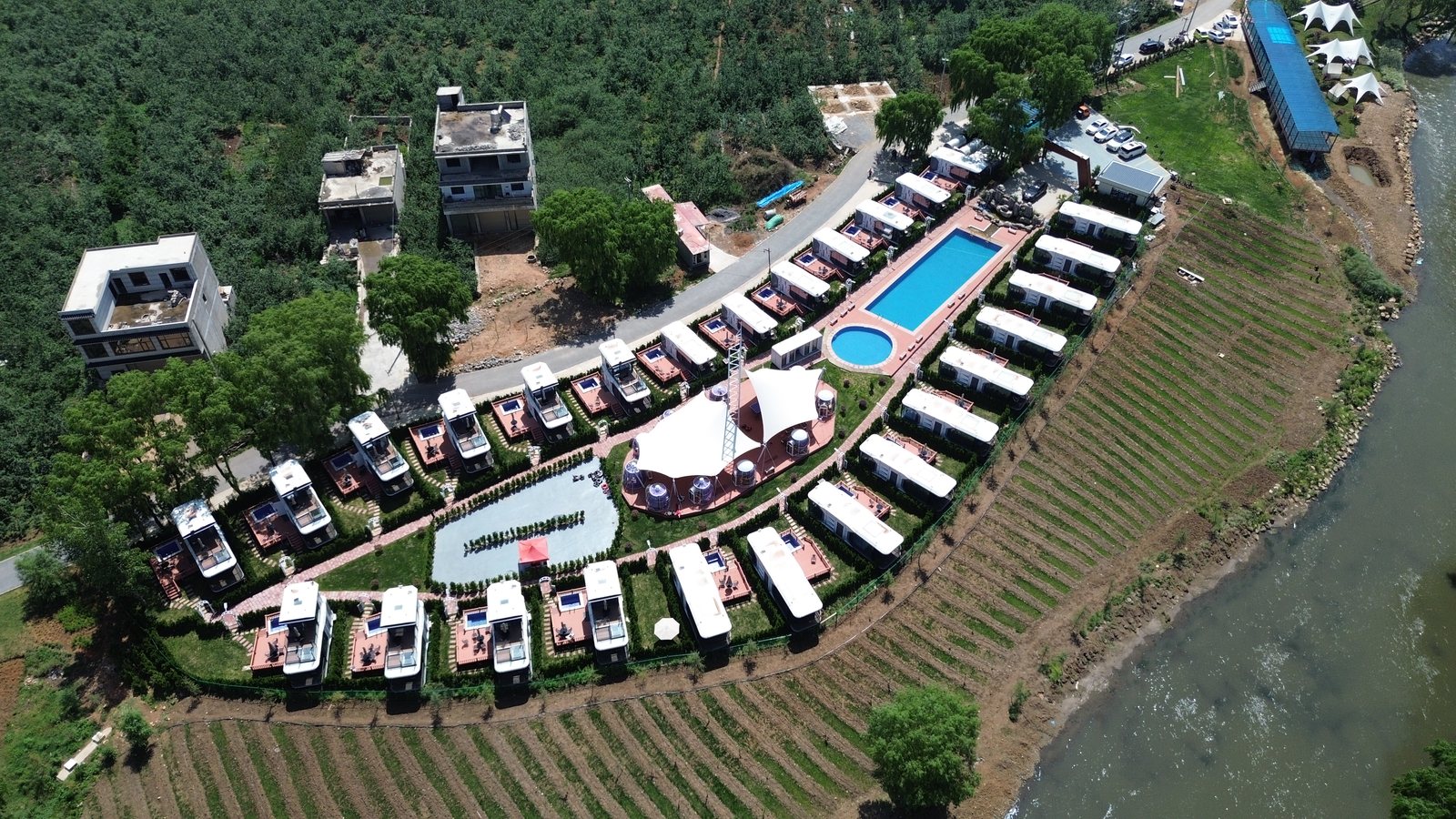 Aerial view of a large capsule cabin resort around a pool beside farmland and water